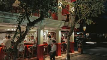 Un área de comedor al aire libre con mesas y sillas, un árbol y un edificio con detalles arquitectónicos en rojo y blanco.