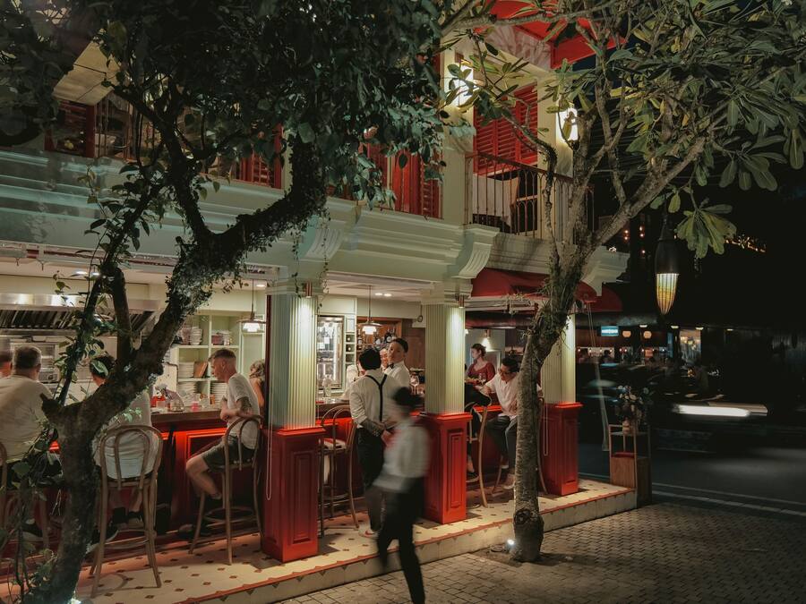 Un área de comedor al aire libre con mesas y sillas, un árbol y un edificio con detalles arquitectónicos en rojo y blanco.
