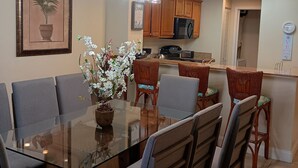 Comedor con mesa de vidrio, seis sillas, un jarrón con flores y una cocina al fondo.
