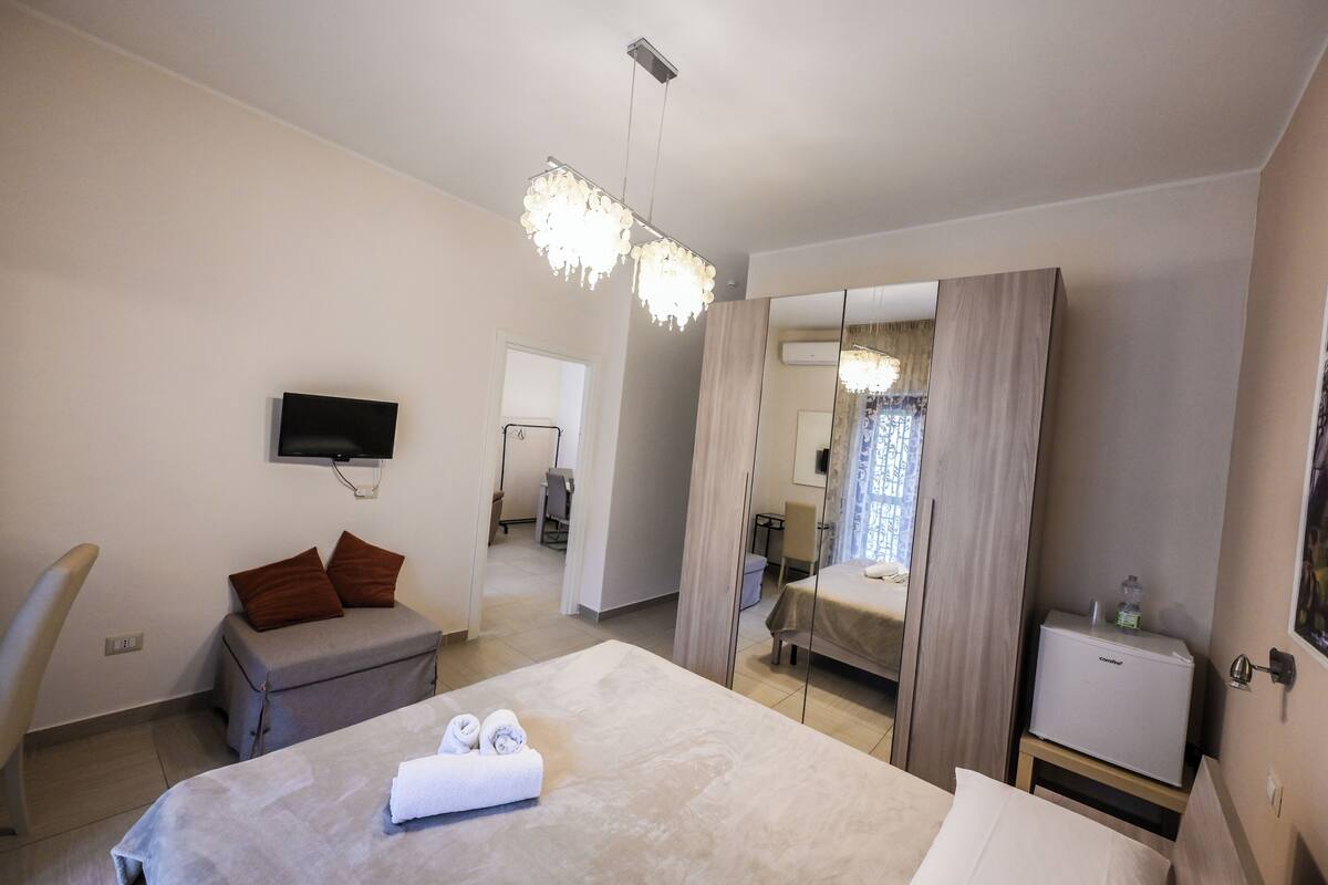 A hotel room with a bed, a seating area, a TV, a minibar, and a mirror.