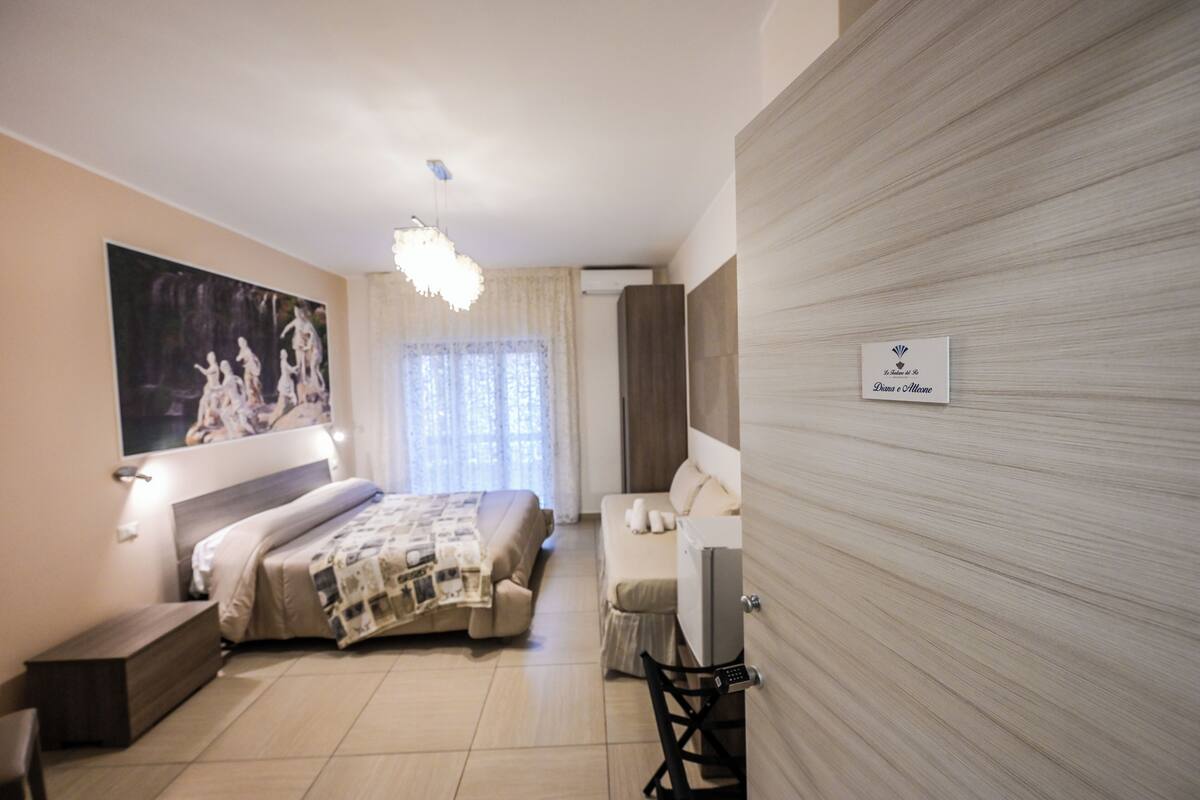 A modern hotel room with a bed, a sofa, a desk, and a large painting on the wall.