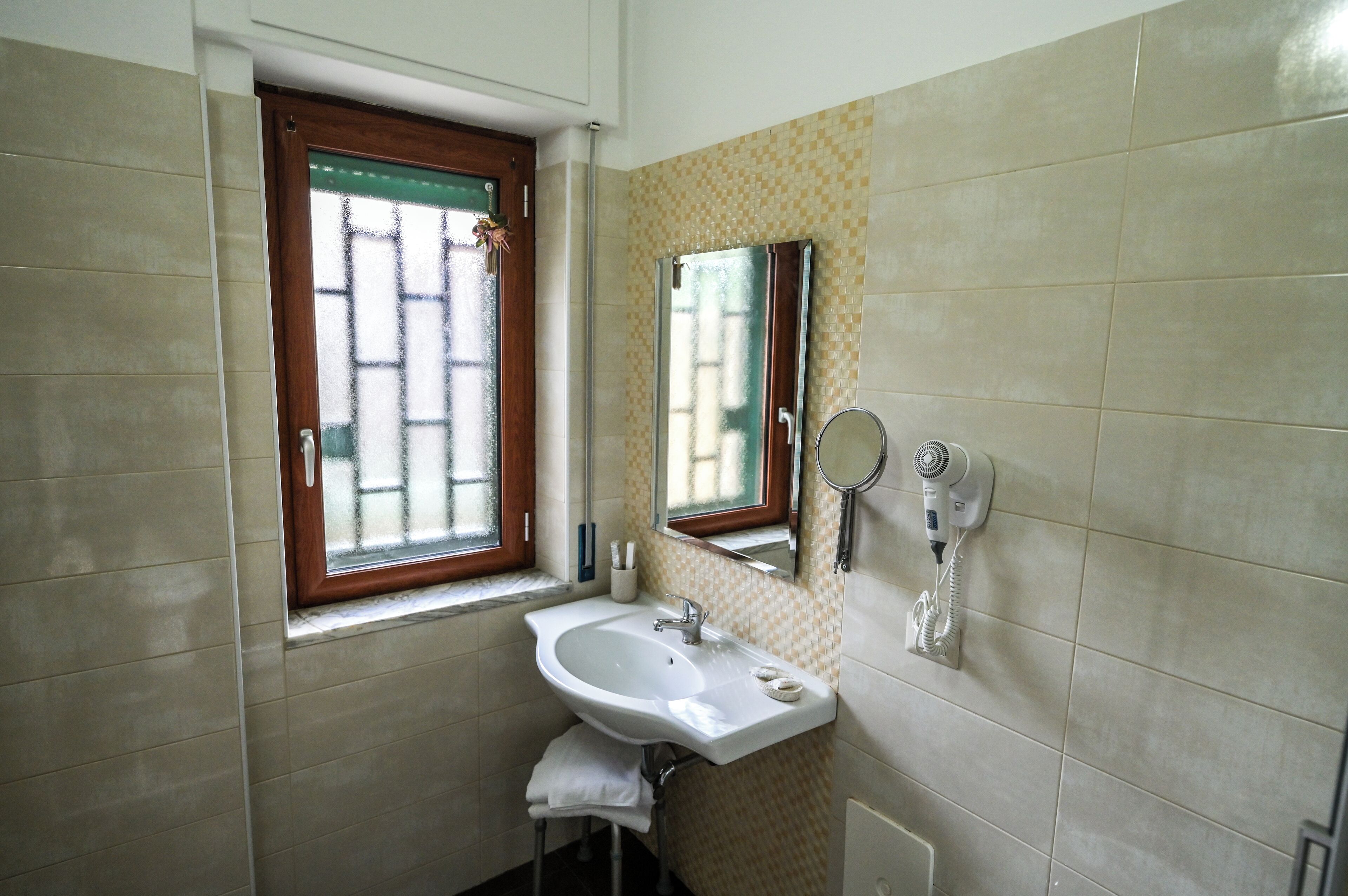 A bathroom with a sink, a window with a wooden frame, a mirror, and a hairdryer on the wall.
