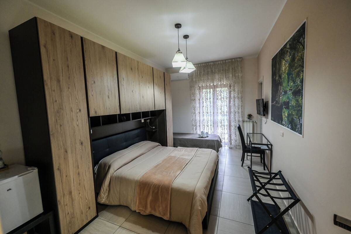 A bedroom with a bed, wooden wardrobe, a desk with a chair, and a painting on the wall.