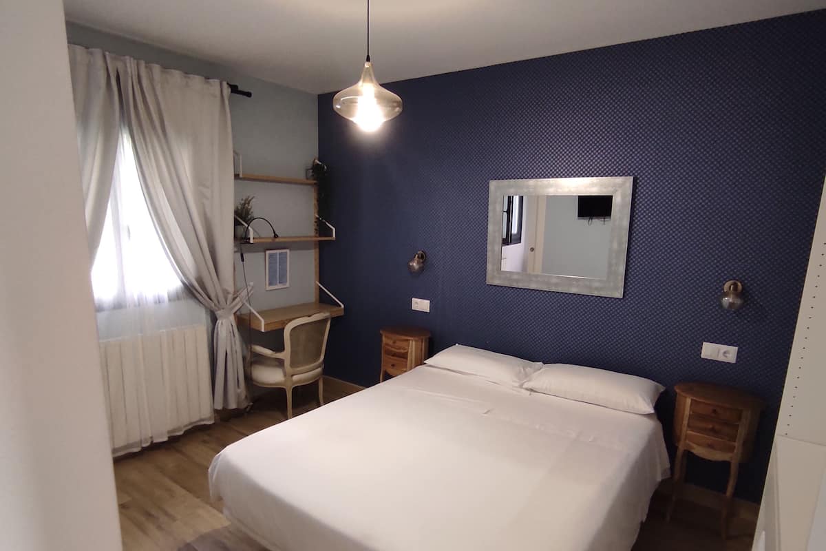 Ein Schlafzimmer mit Bett, Schreibtisch, Stuhl, Pendelleuchte und Spiegel an der Wand.