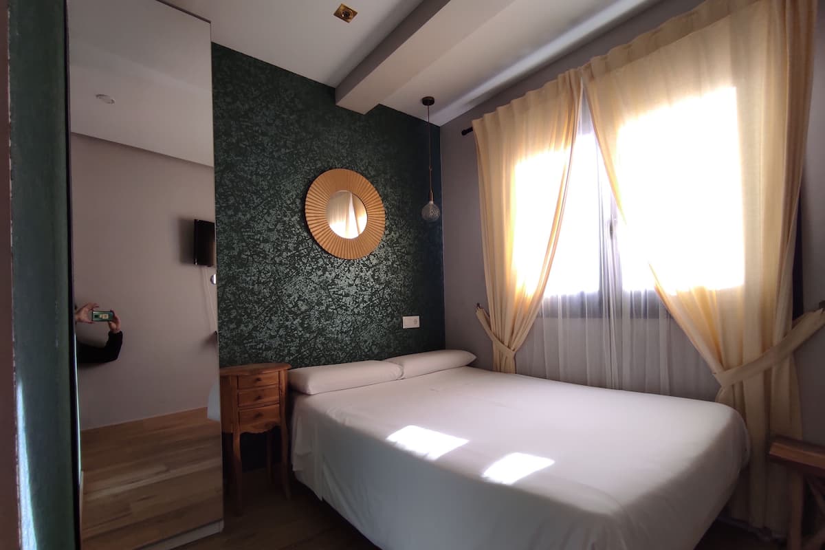 Ein Schlafzimmer mit einem Bett, einem Nachttisch aus Holz, einem runden Spiegel und einem an der Wand befestigten Fernseher.