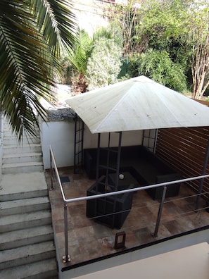 A covered outdoor seating area with a metal railing, a staircase leading up, and a view of the surrounding greenery.