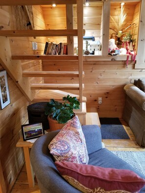 Interior acogedor de una cabaña con escalera de madera, un sofá con cojines estampados, una estantería y una planta en maceta.