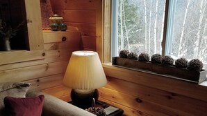 Interior acogedor de una cabaña con revestimiento de madera, un sofá con cojines, una mesita con una lámpara y una ventana con vistas a árboles.