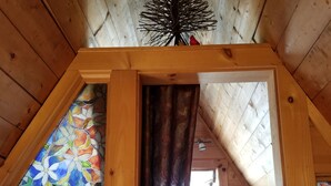 Un dormitorio acogedor en el ático, con techo inclinado, cama con ropa de cama azul y morada, y una ventana con vitrales y motivos florales.