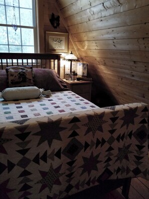 A cozy bedroom with a wooden sloped ceiling, a bed with a quilt, a nightstand with a lamp, and a window with a view of trees. - Cozy, Charming Vermont Chalet close to 5 major ski areas sleeps 4 (Stockbridge)