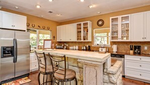 A modern kitchen with white cabinets, a stainless steel refrigerator, a stone island with bar stools, and various kitchen appliances. - FALL FLASH SALE-Perfect Getaway with Natural Spring Swimming Hole! (Spicewood)