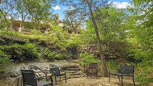A stone patio with wicker furniture, surrounded by lush greenery and a rocky hillside. - FALL FLASH SALE-Perfect Getaway with Natural Spring Swimming Hole! (Spicewood)