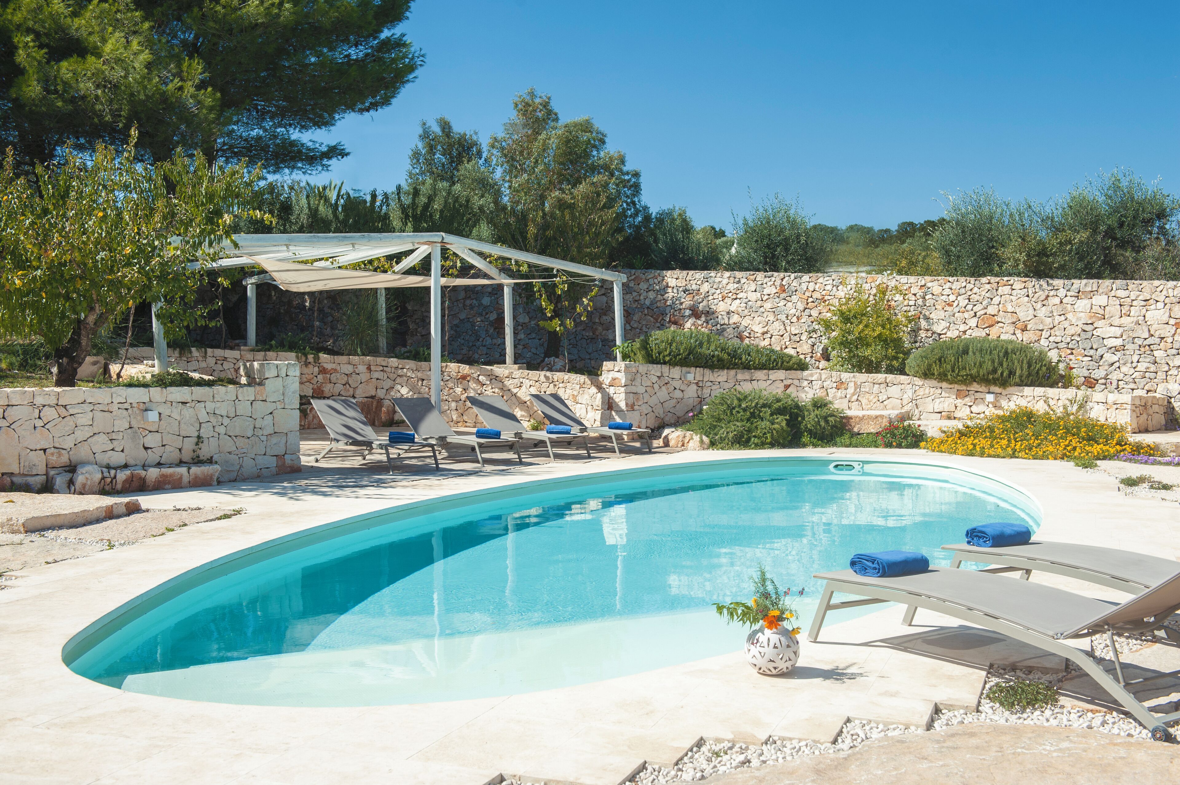 A swimming pool with lounge chairs and a pergola, surrounded by stone walls and greenery.