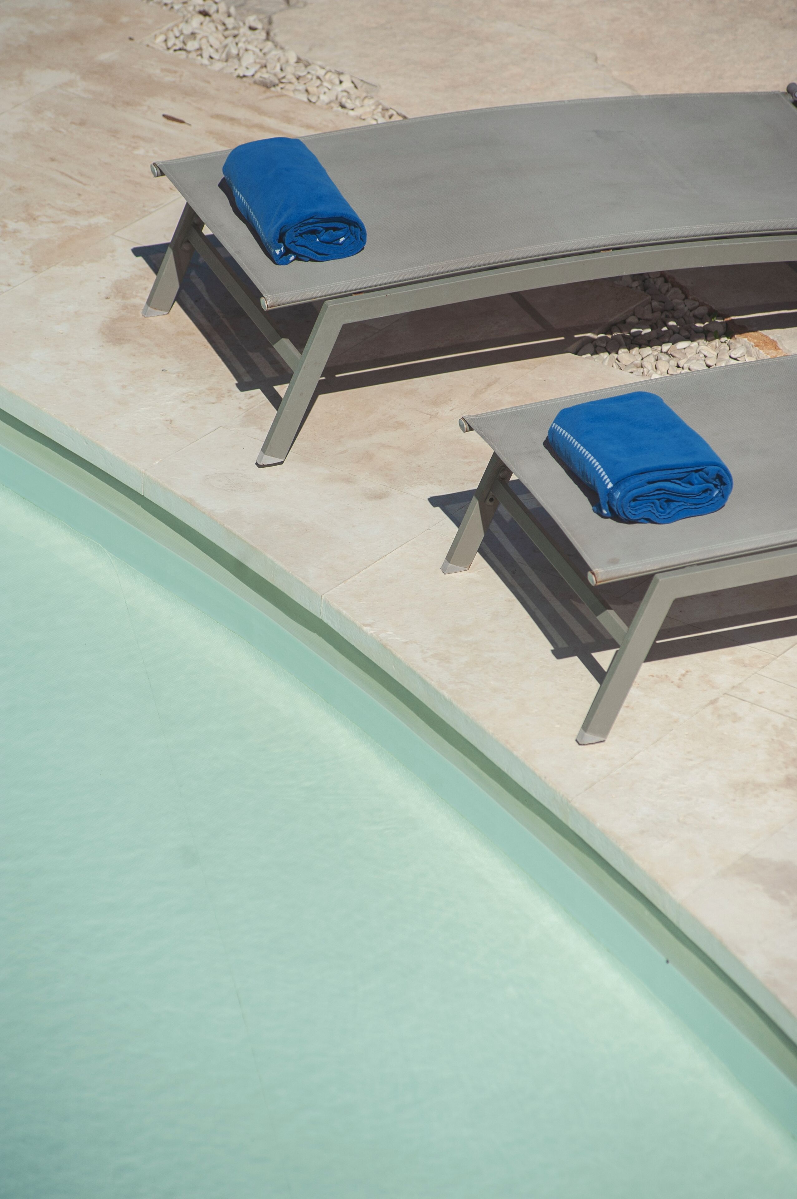 Two sun loungers with blue towels, a pool, and a pebble-lined walkway.