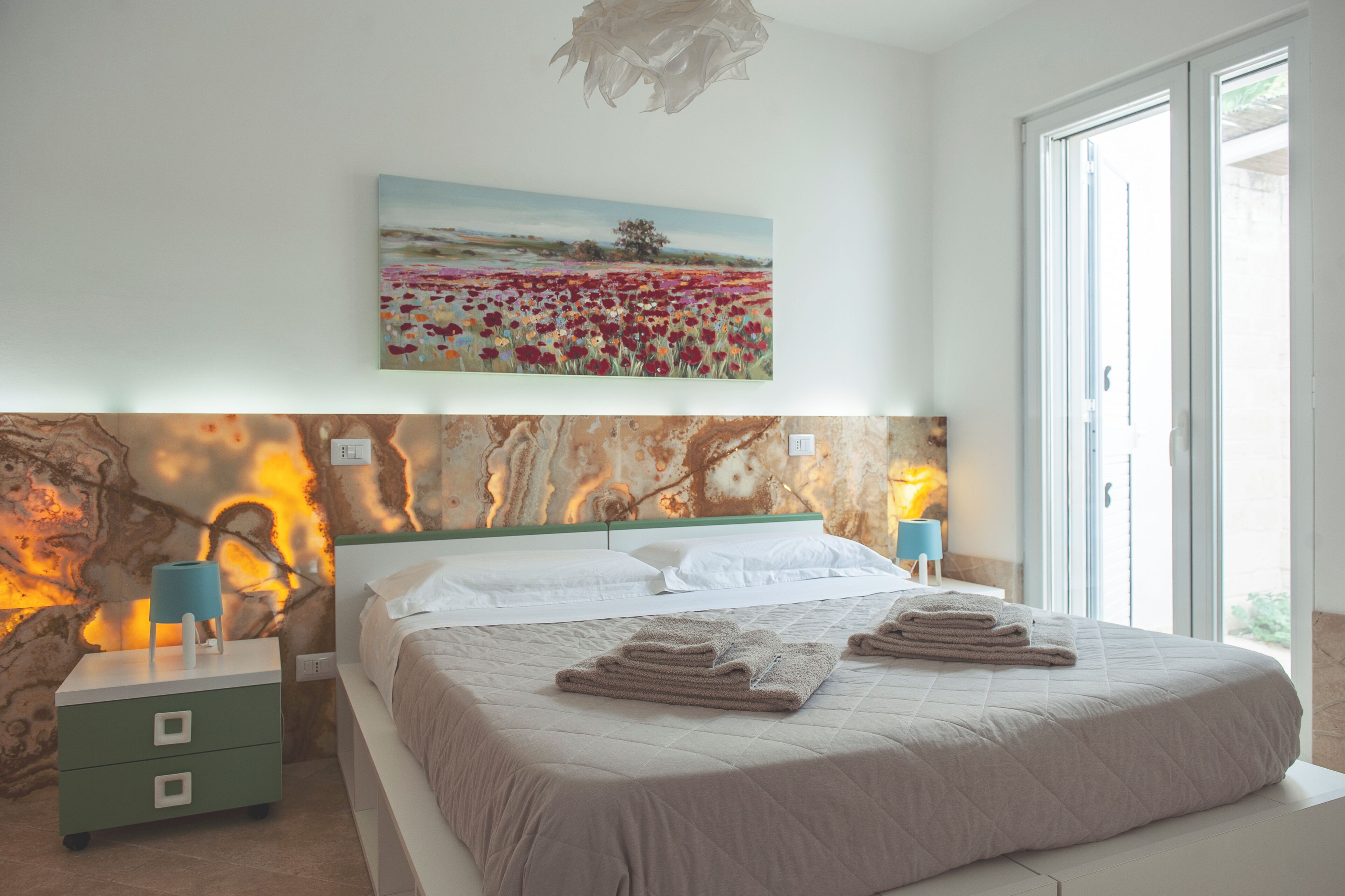 A bedroom with a bed, nightstands, a painting, and a view of the outdoors.