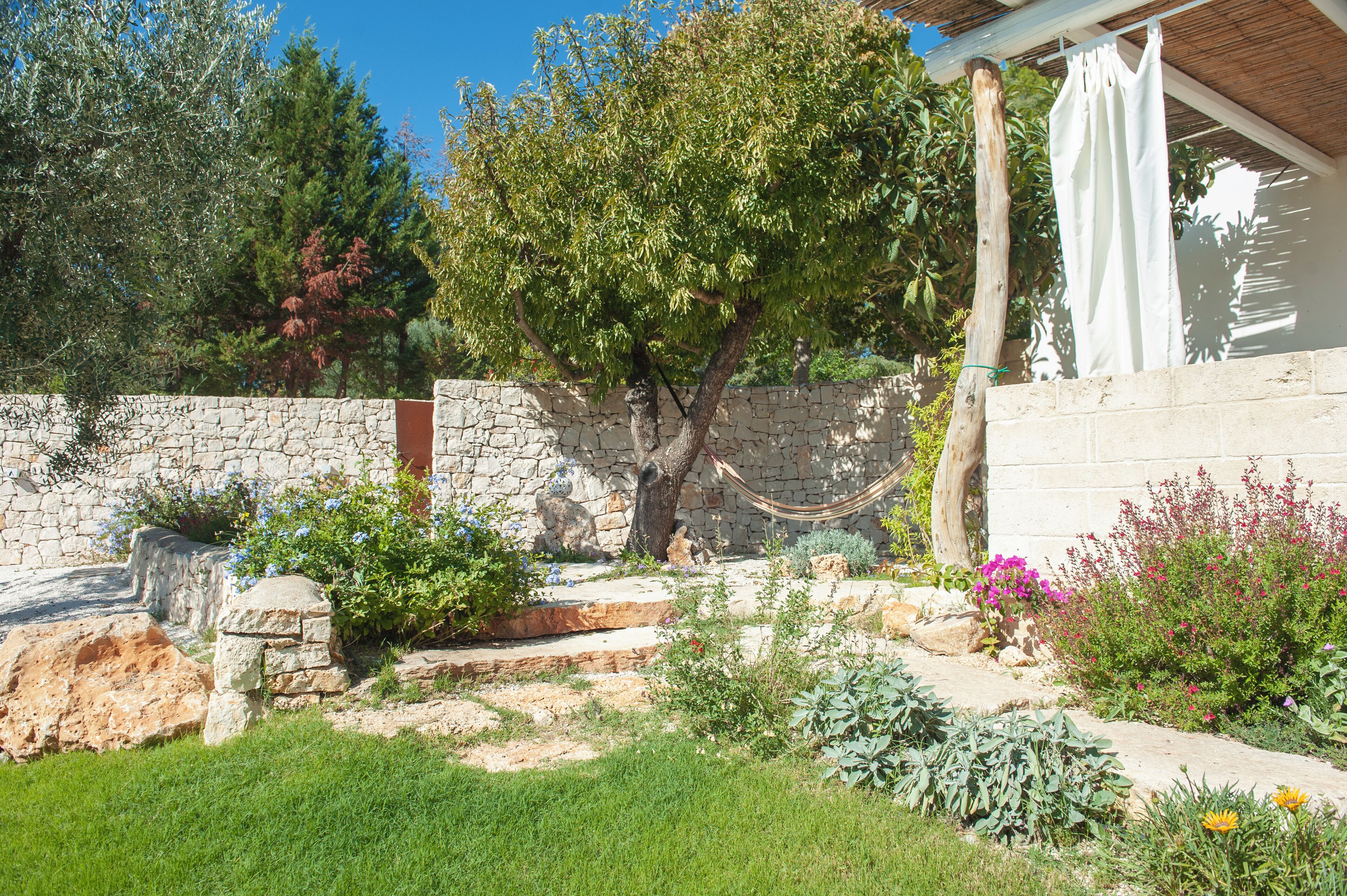 Un mur de pierres, un hamac et une variété de plantes et de fleurs dans un jardin.
