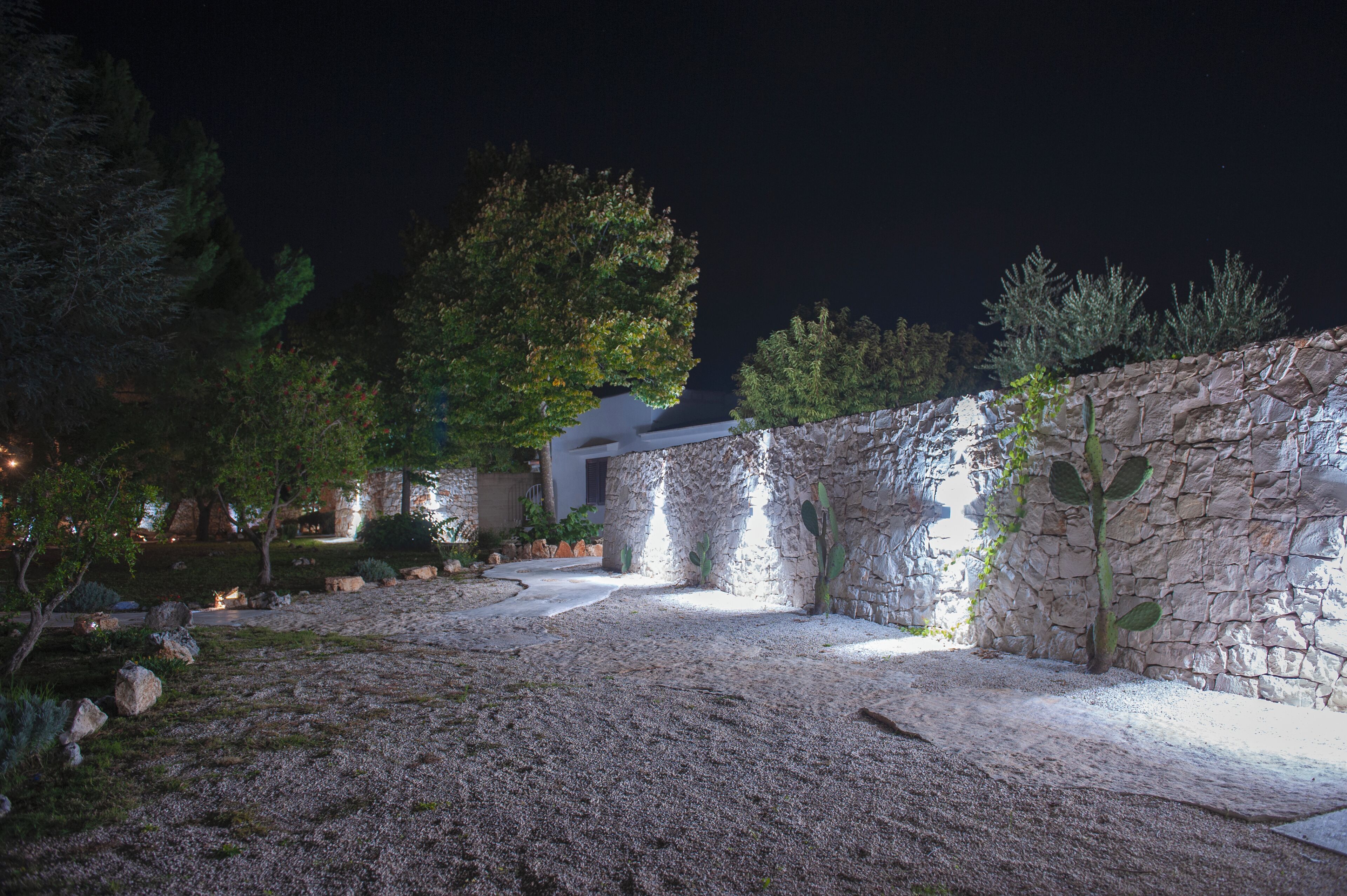 Un espacio exterior nocturno con un muro de piedra, árboles y un sendero que lleva a un edificio.