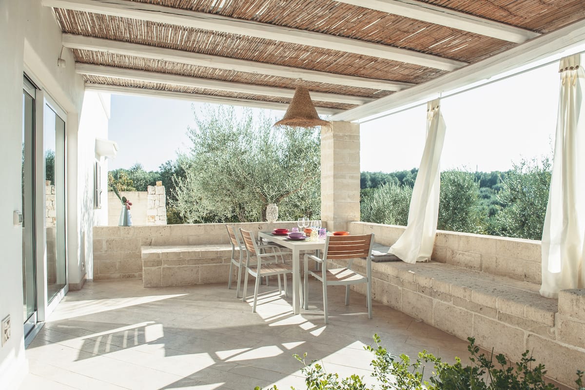 An outdoor patio with a table and chairs, a hanging light fixture, and a view of greenery.