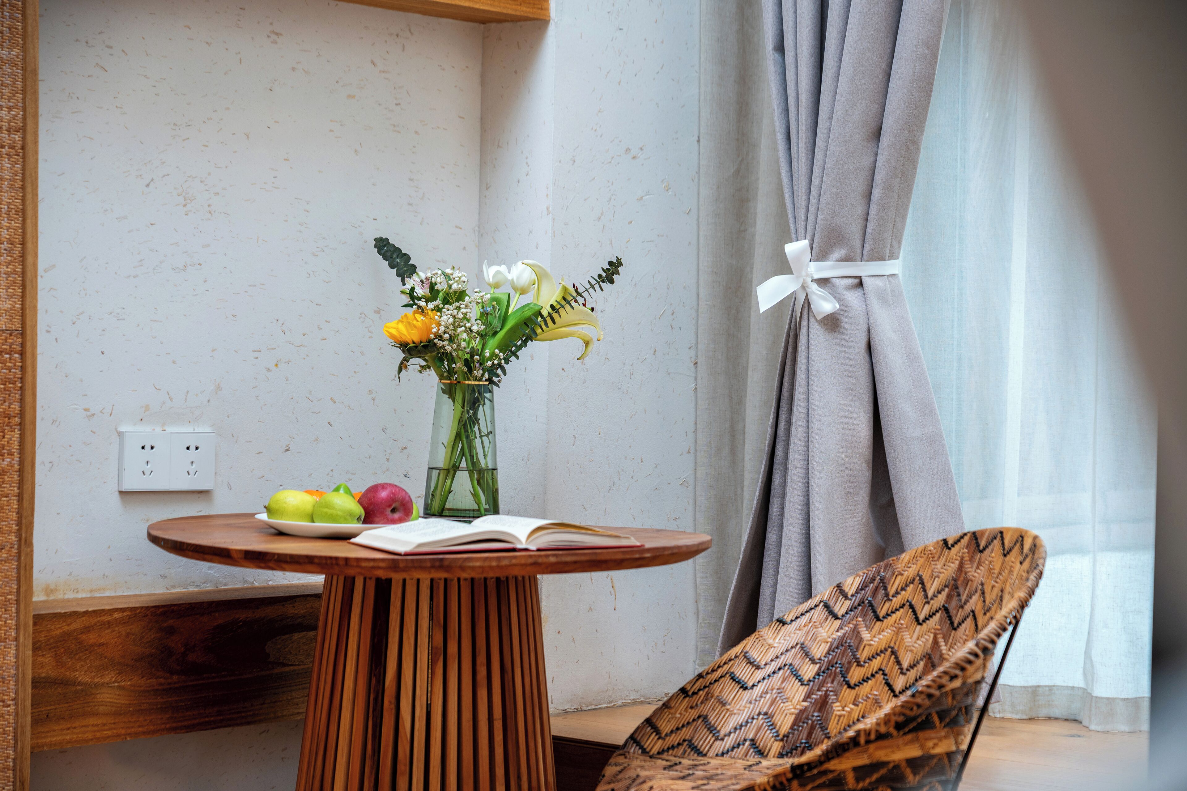A room with a round wooden table, a wicker chair, a vase with flowers, and a book.