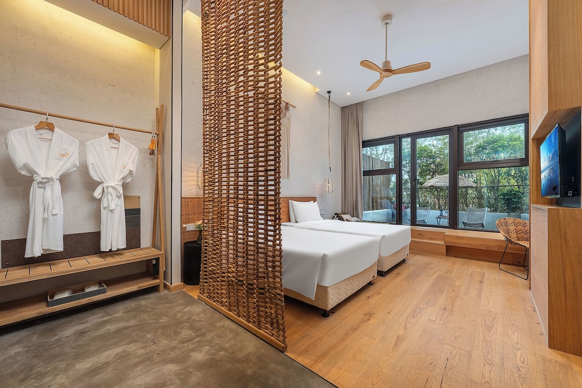 Habitación de hotel moderna con una cama grande, una zona de estar, un televisor y un balcón con vistas a árboles.