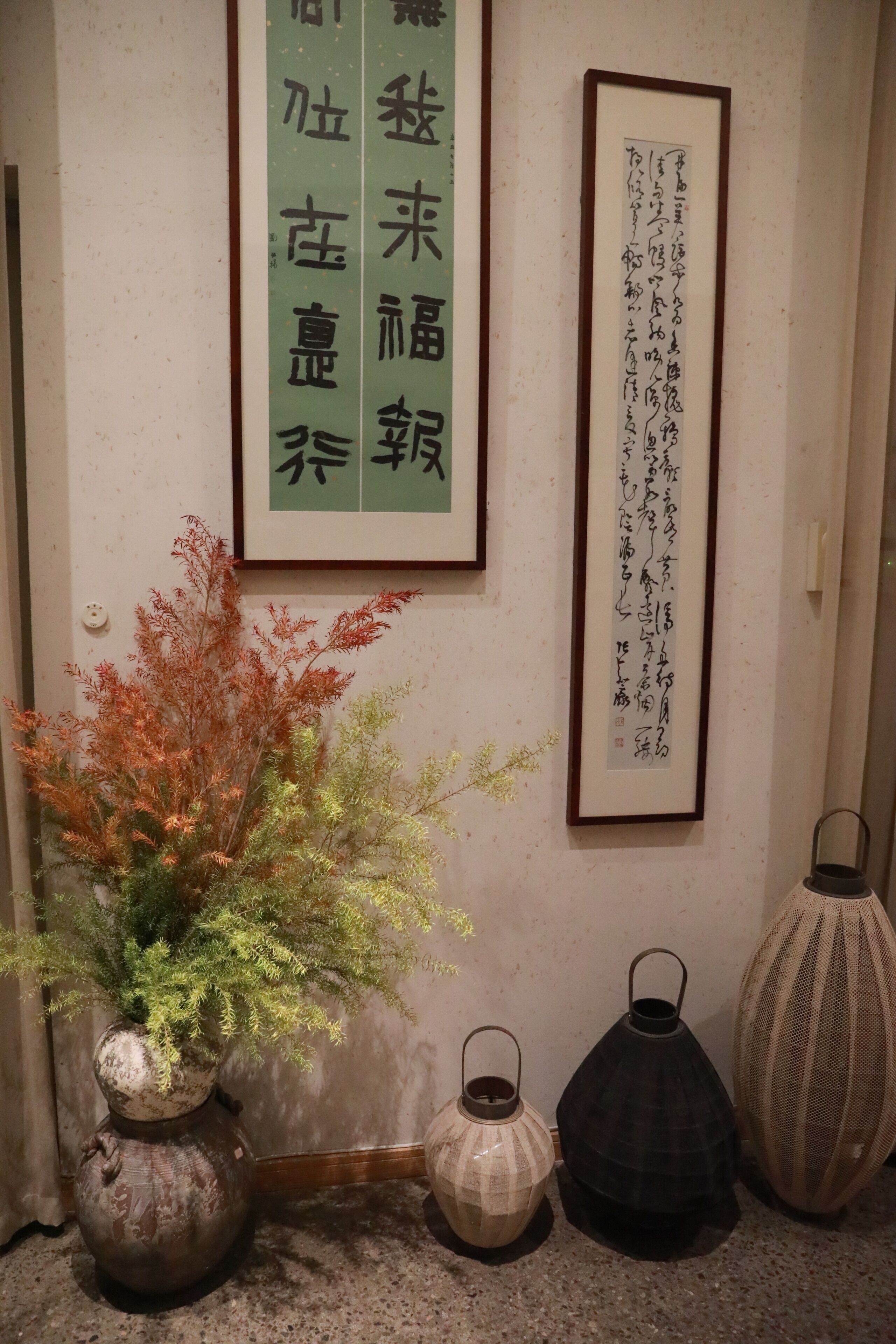 A room with a traditional Japanese aesthetic, featuring a framed calligraphy piece on the wall, a potted plant, and three decorative lanterns.
