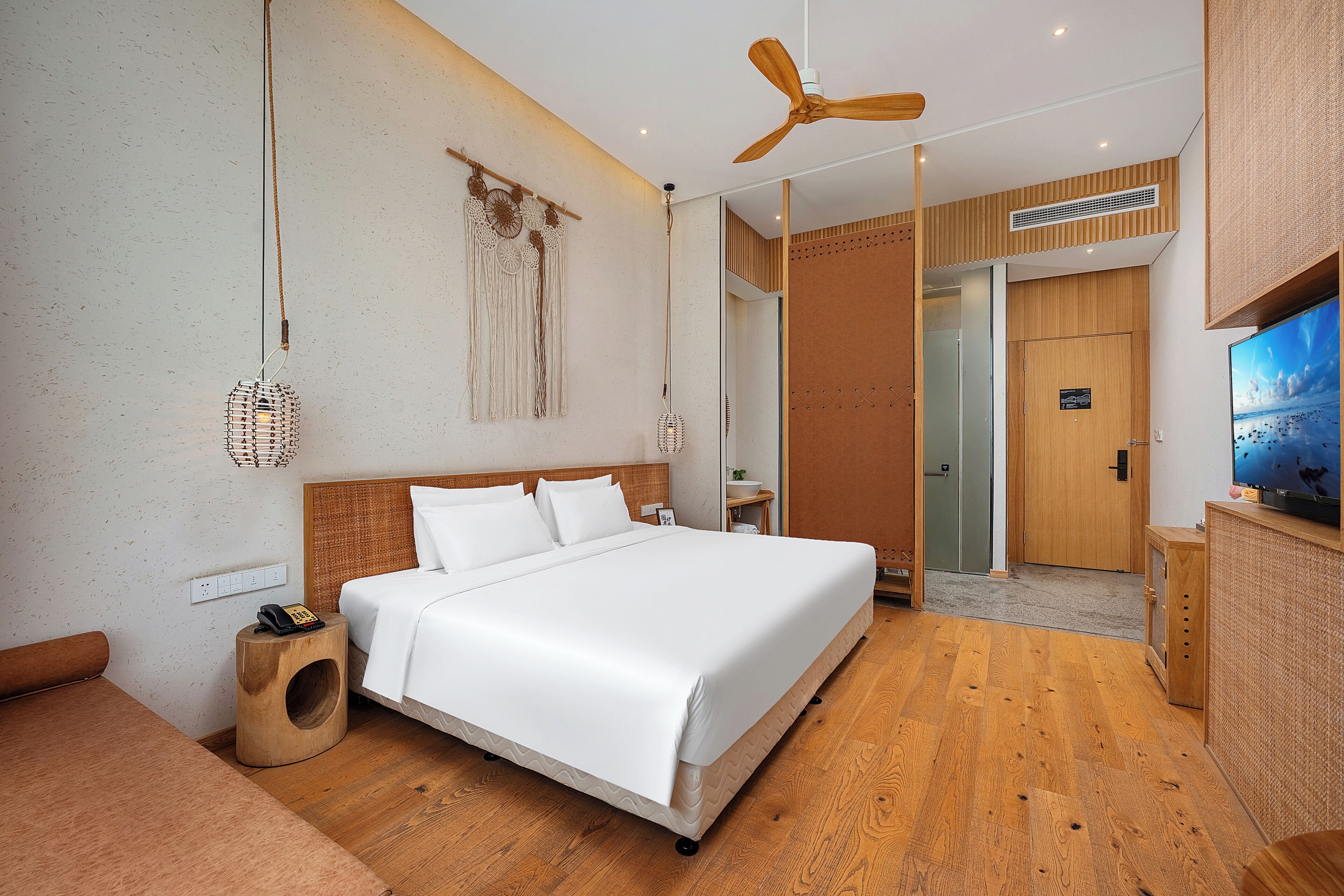 Una habitación de hotel moderna con una cama grande, piso de madera, ventilador de techo, televisor montado en la pared y una mesita de madera.
