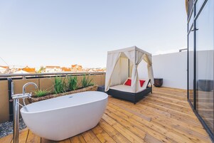 A modern outdoor bathroom with a freestanding bathtub, a cushioned seating area with a canopy, and a wooden deck.
