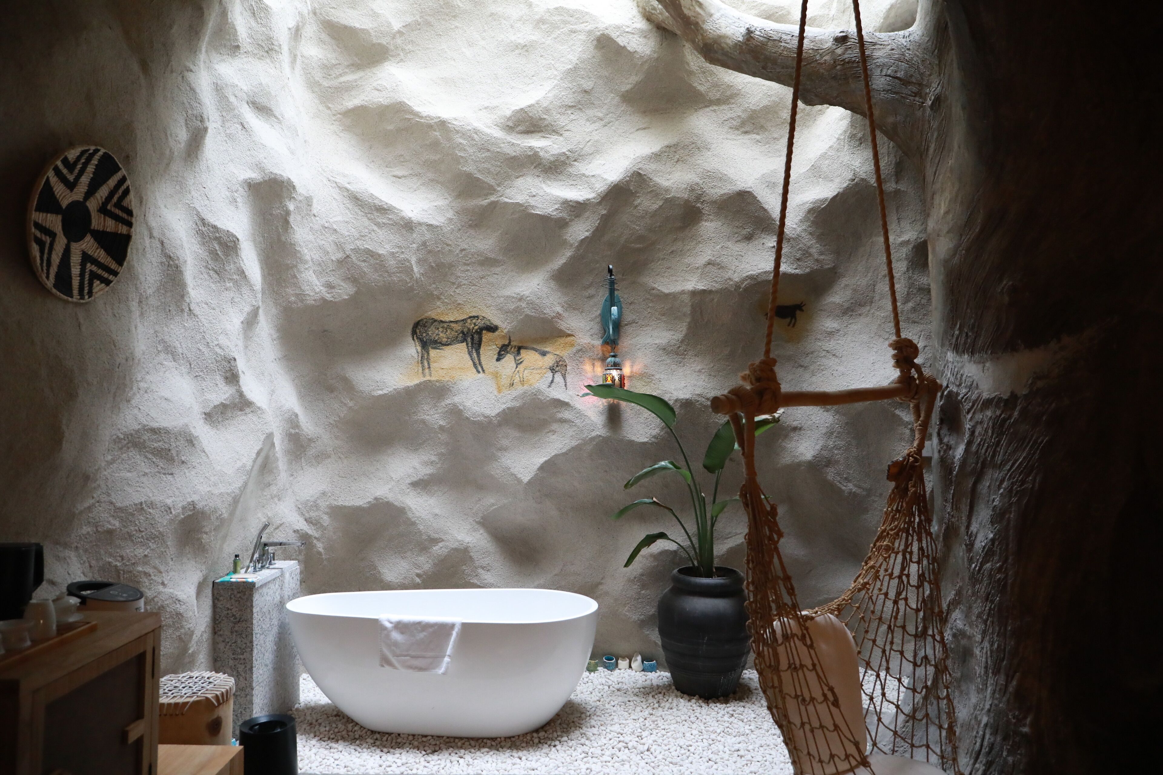 A bathroom with a white bathtub, a stone wall with animal paintings, a hanging macrame swing, and a potted plant.