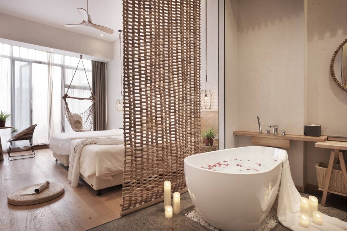 Un dormitorio moderno con una cama grande, una silla colgante, un separador tejido, una bañera con pétalos de rosa y un tocador de madera.