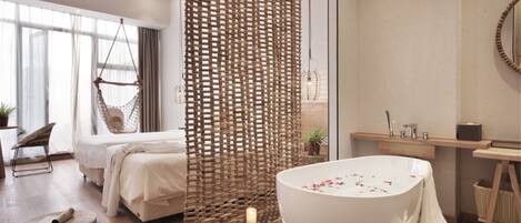 A modern bedroom with a large bed, a hanging chair, a woven partition, a bathtub with rose petals, and a wooden vanity.