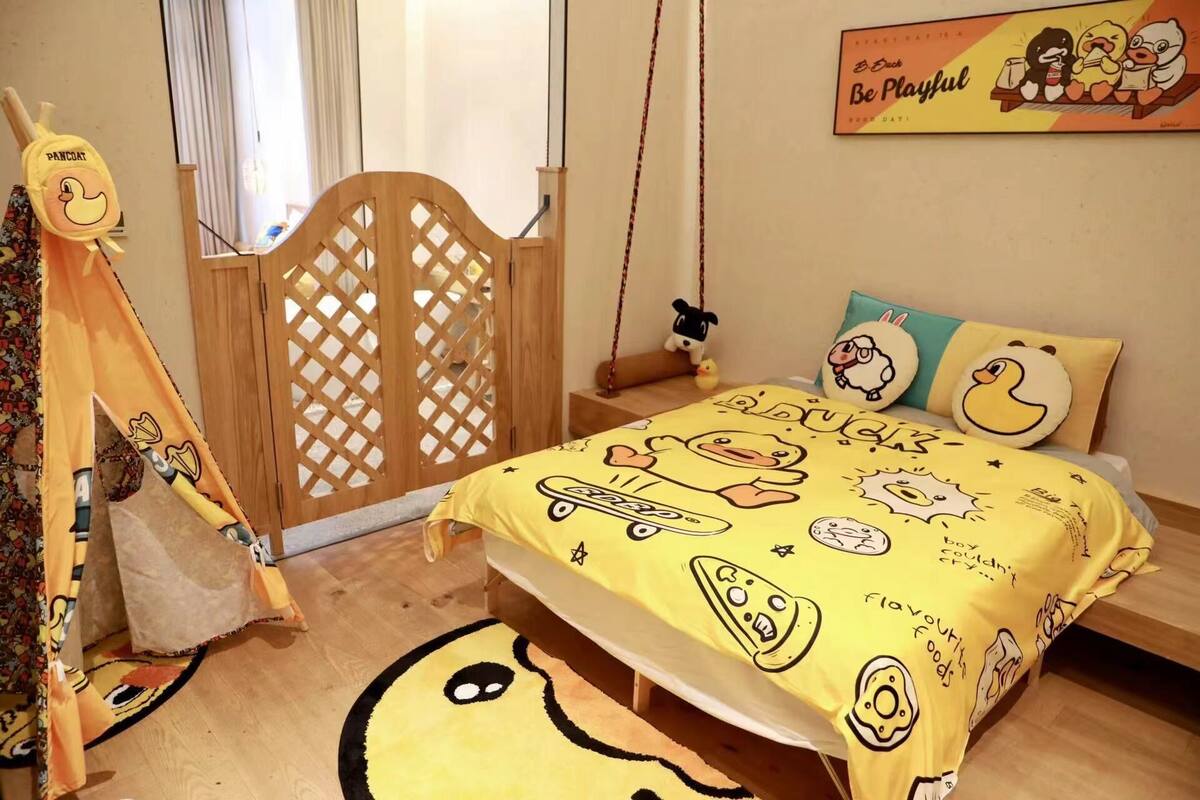 Un dormitorio con una cama cubierta con un edredón amarillo con personajes de dibujos animados, un armario de madera, una mesita de noche con una lámpara y una pared con un póster.
