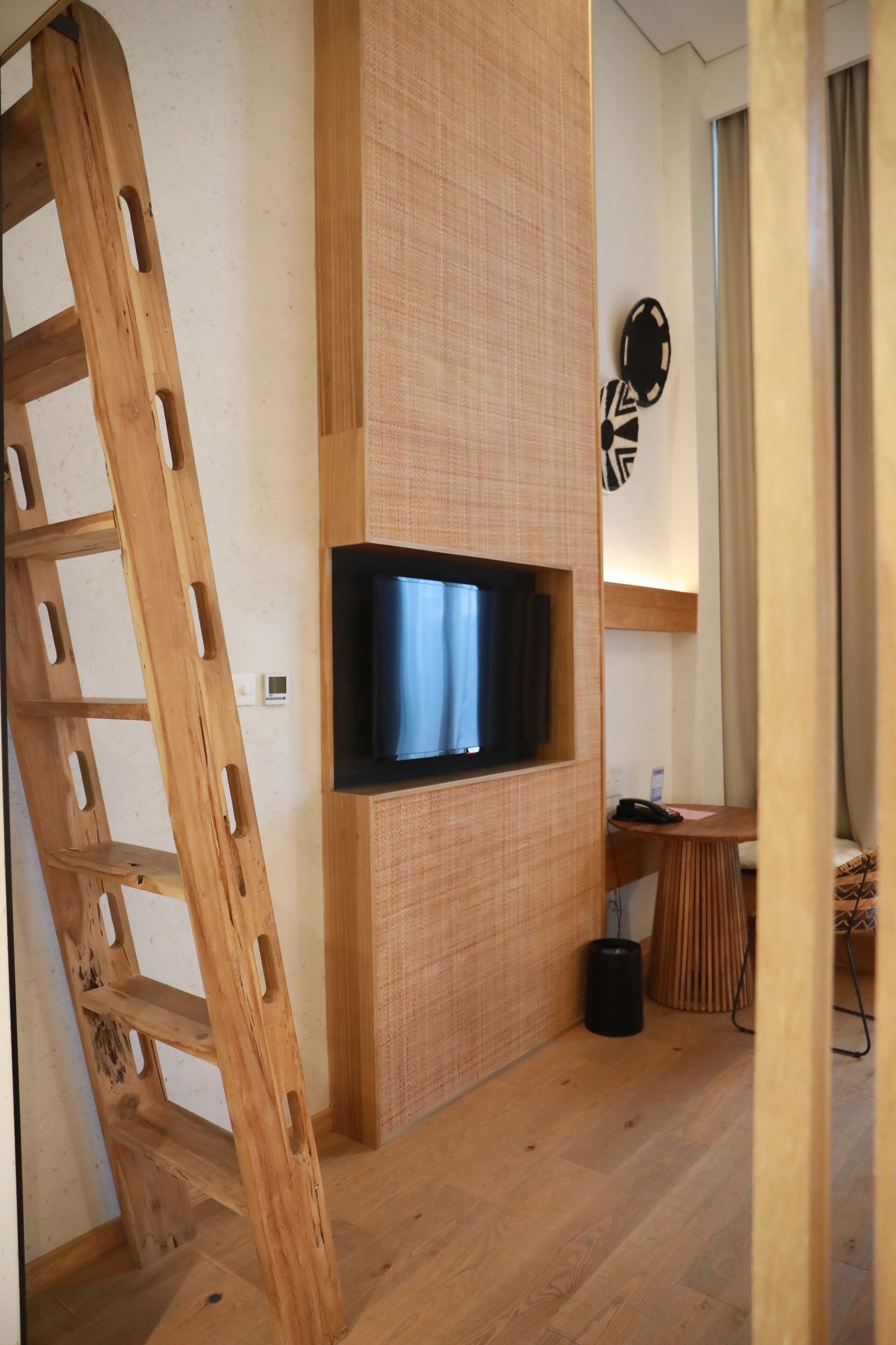 A room with a wooden ladder, a flat-screen TV mounted on a textured wall, a small table with a telephone, and a trash bin.