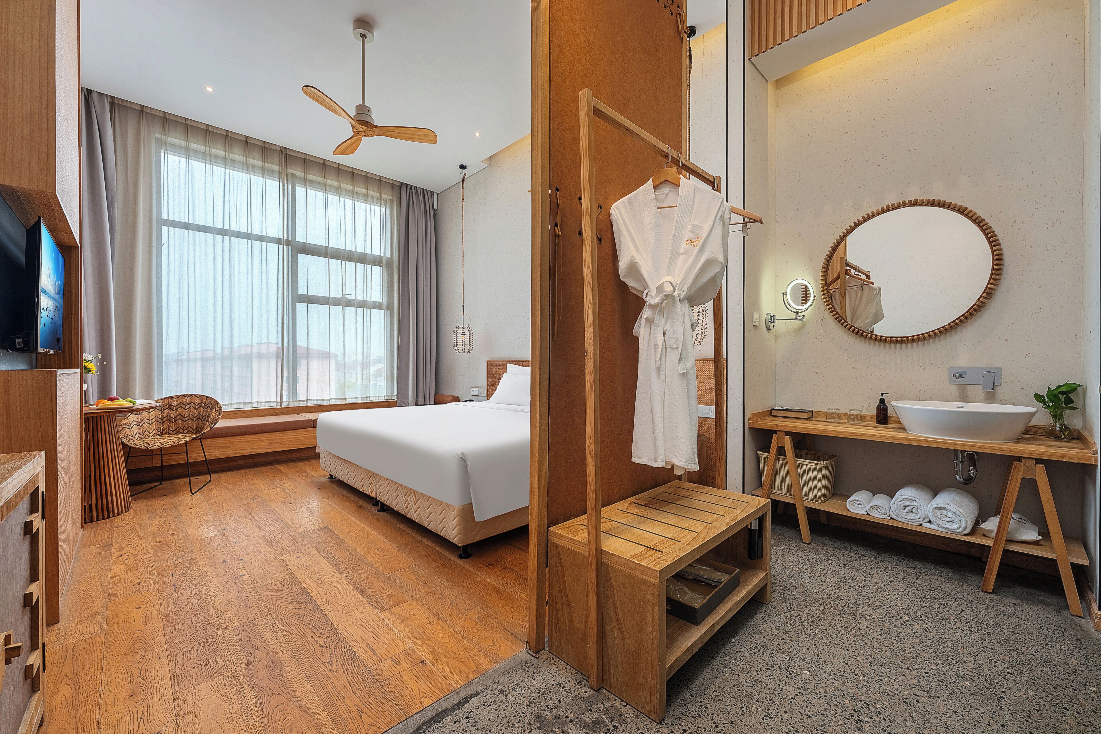 A modern hotel room with a large bed, a wooden bench, a TV, a ceiling fan, and a bathroom with a sink and mirror.