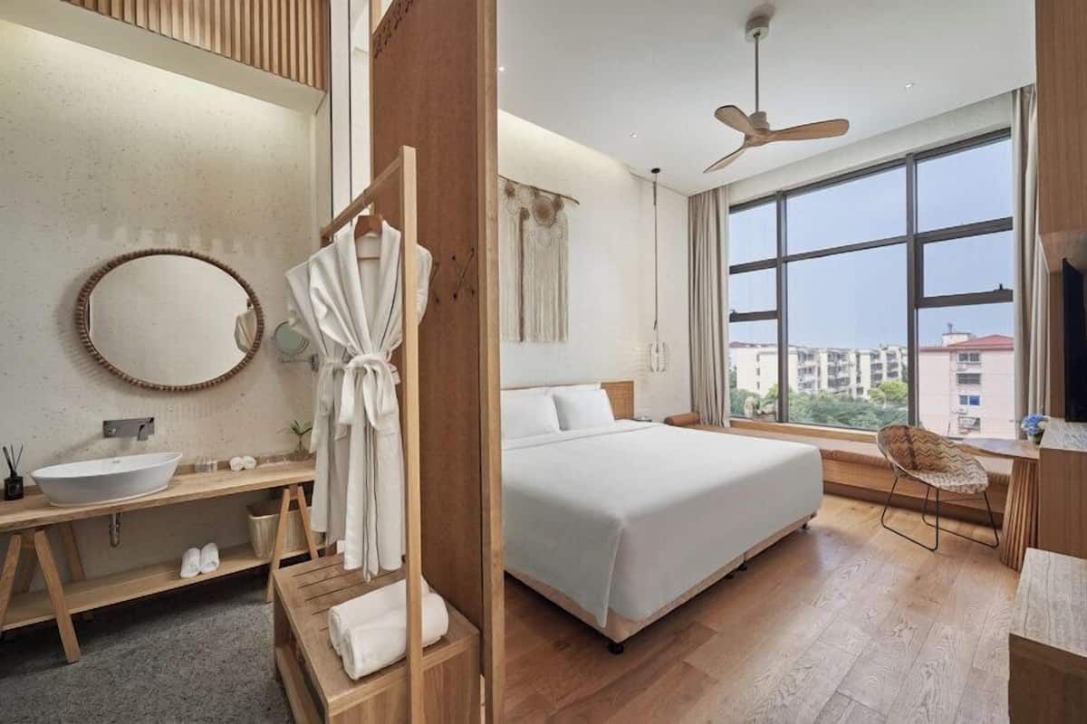 Habitación de hotel moderna con una cama grande, un tocador de madera, un espejo redondo y un ventanal con vistas a la ciudad.