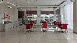A spacious dining area with red chairs and tables, a chandelier, and a modern kitchen in the background. - Kanasha Islami (Medan)