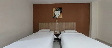 A hotel room with two single beds, a wooden headboard, a painting of flowers, and a bedside table with a phone and a lamp.