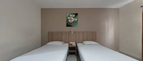 A hotel room with two single beds, a wooden headboard, a small table with a phone, and a painting of flowers on the wall.
