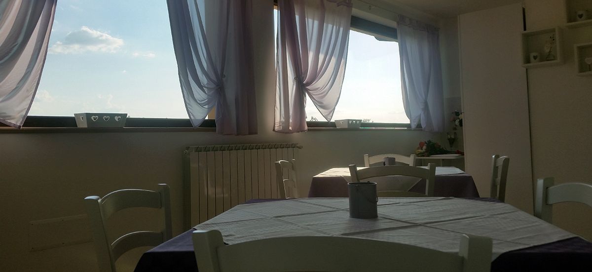 A dining area with a table set for four, white chairs, a window with curtains, and a radiator below.