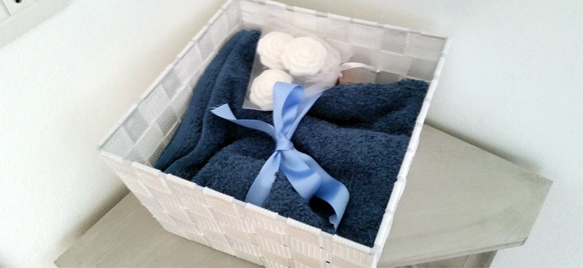 Un cesto bianco con un nastro blu, contenente asciugamani arrotolati e una bottiglia, su una superficie di legno.