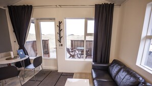 Une pièce comprenant un canapé en cuir noir, une table basse blanche, une table à manger avec des chaises, et donnant sur une terrasse en bois et une plage.