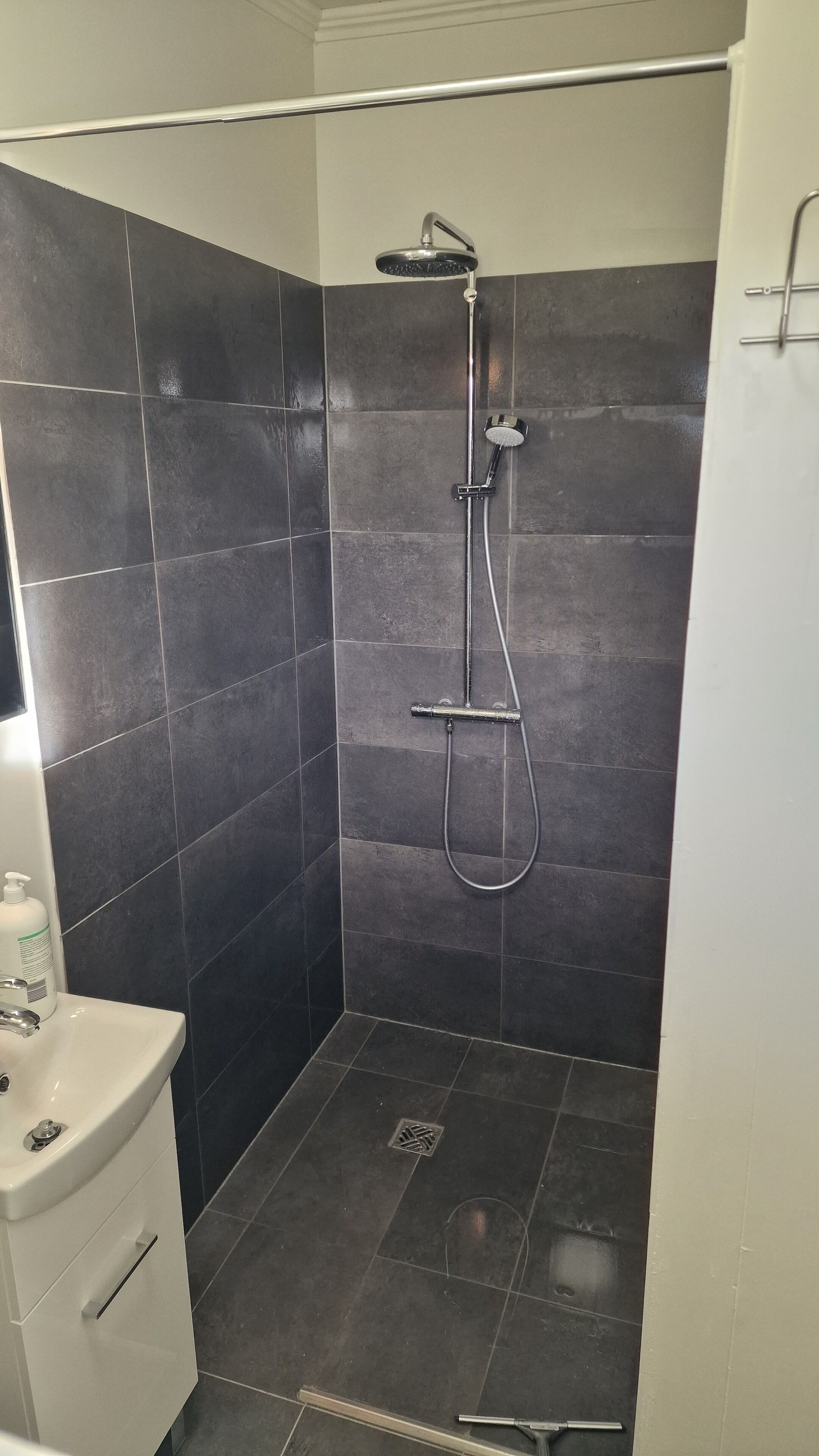Ducha con cabezal de ducha de mano y grifo empotrado, lavamanos blanco con dispensador de jabón y paredes de azulejos oscuros.