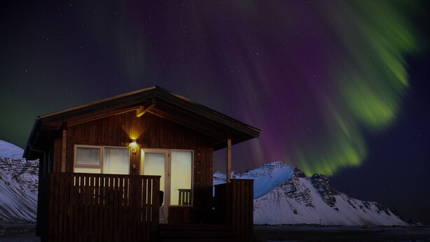Une cabane en bois dotée d’une véranda et d’une fenêtre illuminée, se dressant sur fond de montagne enneigée, tandis que les aurores boréales illuminent le ciel.