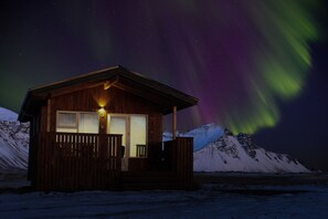 A wooden cabin with a porch and a lit window, set against a snowy mountain backdrop with the Northern Lights in the sky. - Aurora Cabins (Höfn)