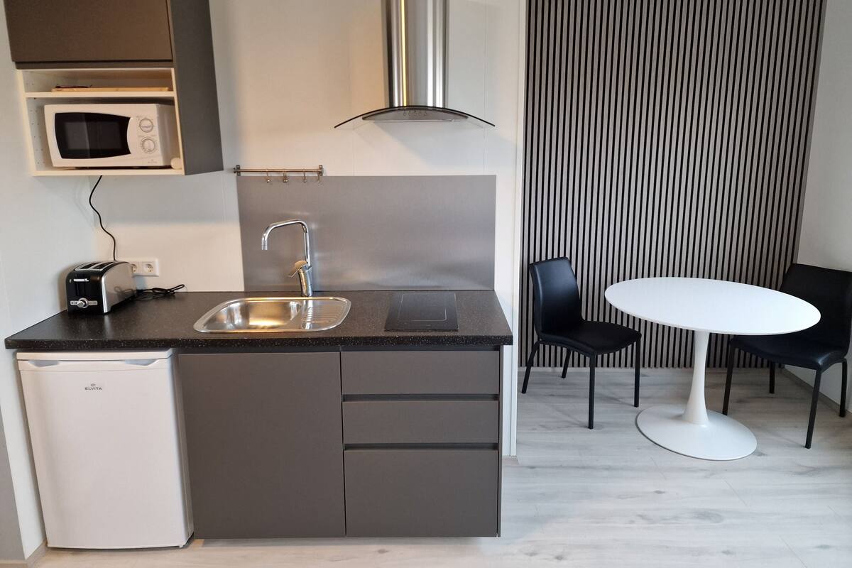 A compact kitchen with a built-in microwave, a stainless steel range hood, a sink, and a compact dishwasher. There is a small dining area with a white table and two black chairs.