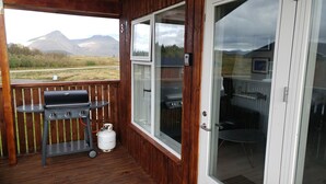 Une terrasse en bois équipée d’un barbecue, d’une bouteille de gaz et d’une porte vitrée donnant sur un espace de repas intérieur avec une table et des chaises.