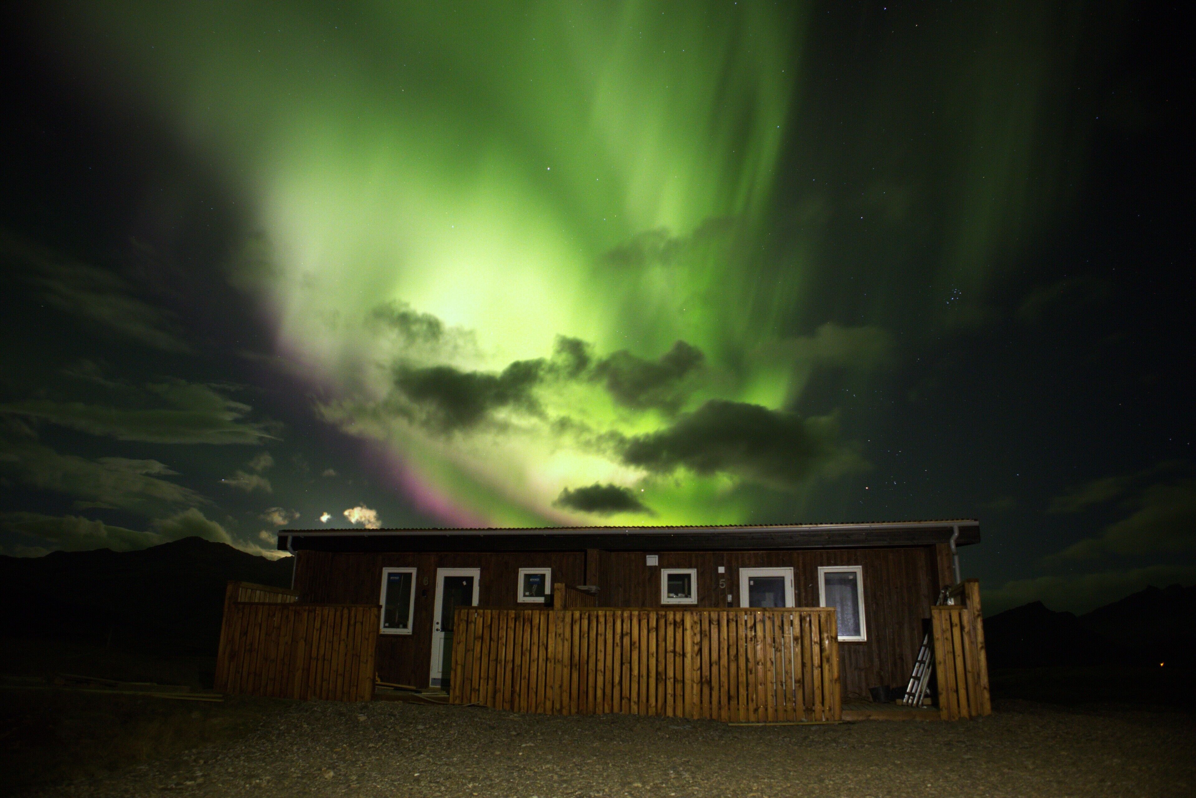A wooden cabin with a single door and windows, surrounded by a wooden fence, under a night sky with visible aurora borealis.