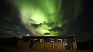 A wooden cabin with a single door and windows, surrounded by a wooden fence, under a night sky with visible aurora borealis.