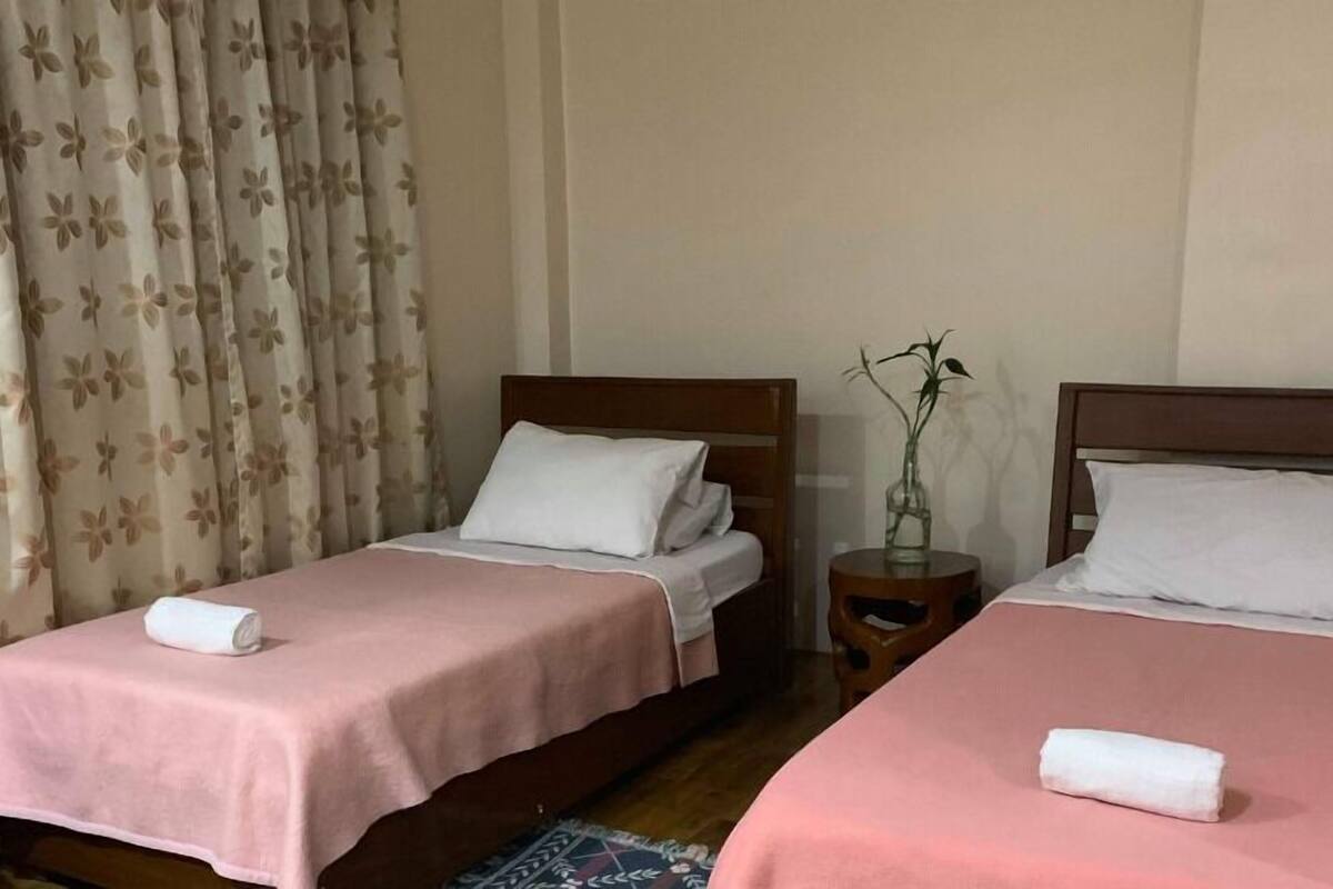 Ein Hotelzimmer mit zwei Einzelbetten, jeweils mit einer rosa Bettdecke und weißen Kissen, einem hölzernen Kopfteil und einem kleinen Nachttisch mit Blumen.'