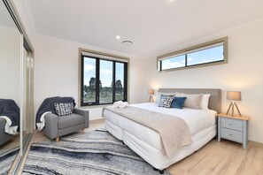 A bedroom with a large bed, a grey sofa, a small bedside table, and a window with a view of trees. - TheHorizon7 (Pokolbin)