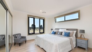 Quarto com uma cama grande, uma cadeira, uma mesinha de cabeceira e uma janela com vista para árvores.